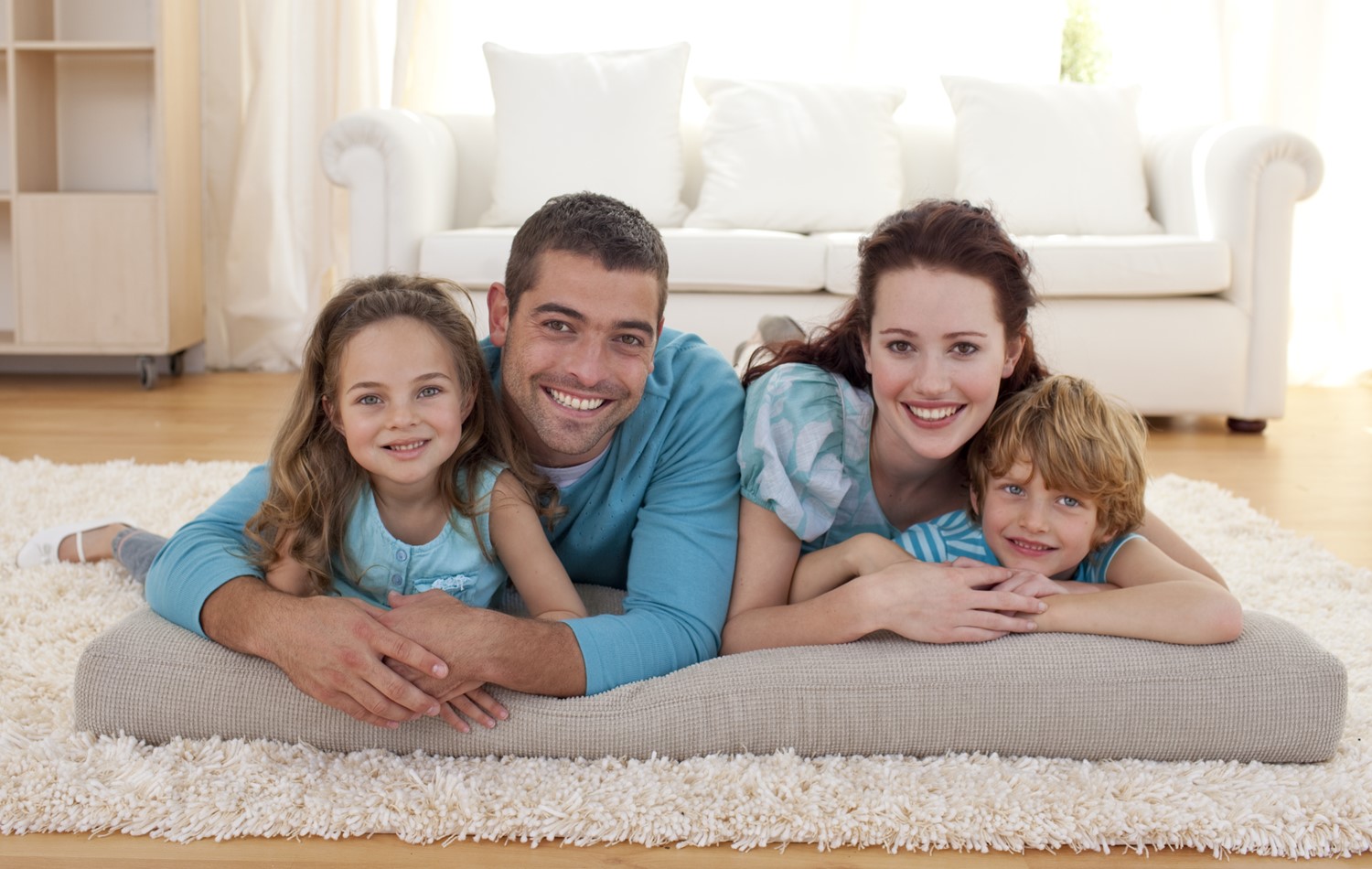 A Smiling Daughter, Dad, Mom, and Son Lay on a Cushion on a Beige Shag Carpet in Front of a Couch