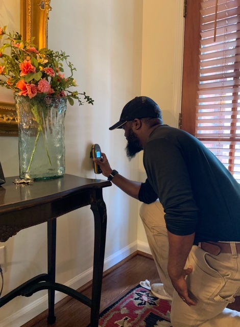  A Green Homes Solutions Technician Kneels and Holds a Device Against a Wall to Inspect for Mold While Another Man Watches