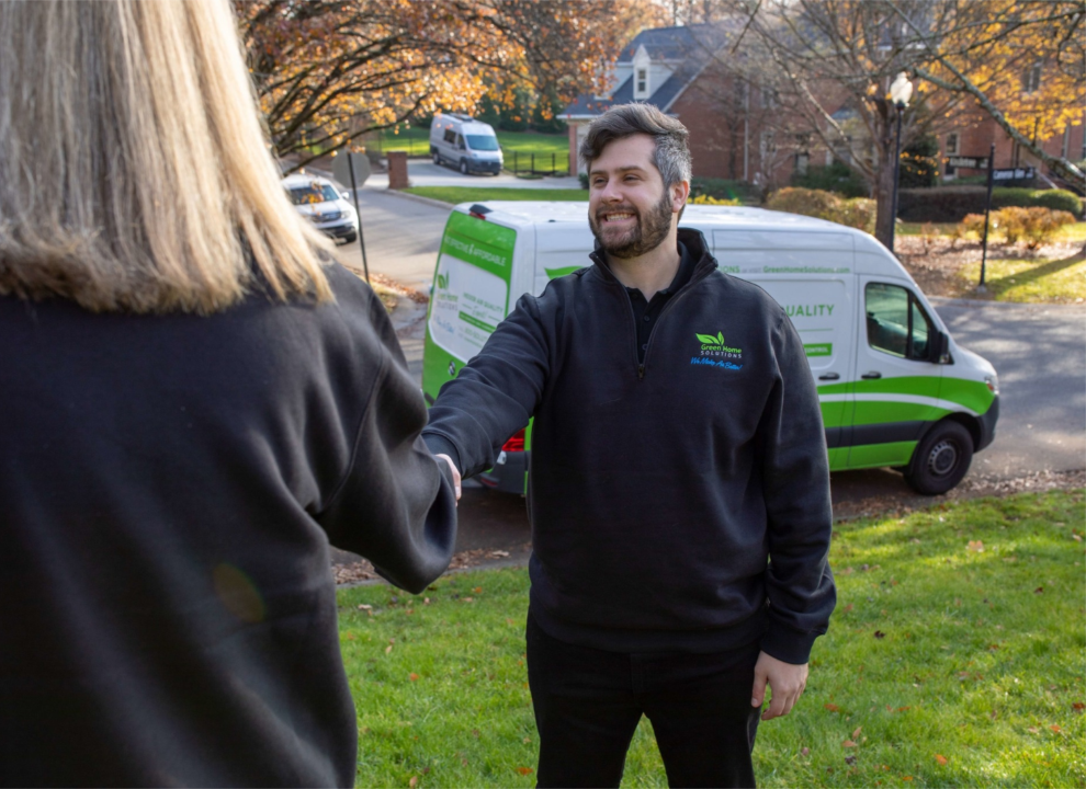 A man in a Green Home Solutions pullover shakes hands with a woman pictured from behind with a Green Home Solutions work van behind him.