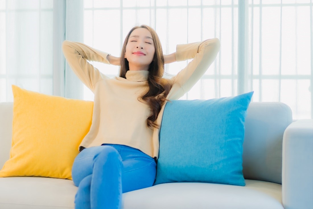 A Woman Sits on a Couch Between a Yellow and a Blue Pillow with Her Hands Behind Her Head and Eyes Closed Taking a Deep Breath