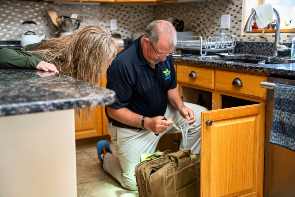 A man in a Green Home Solutions polo kneels on the floor and takes a sample of something from under a kitchen sink while a woman observes.