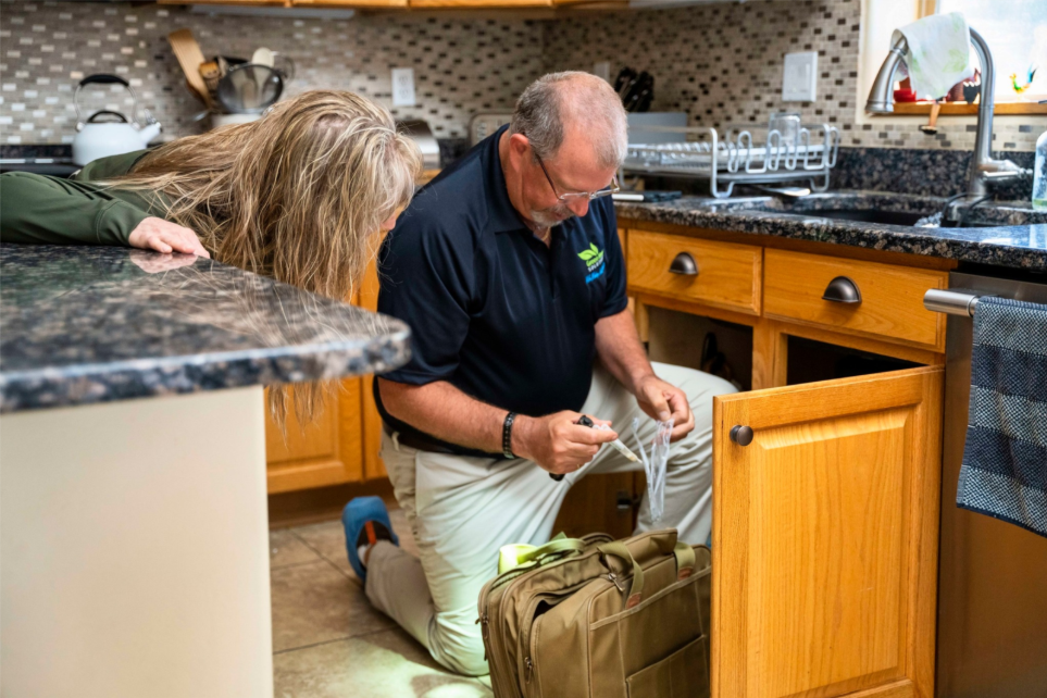 A man in a Green Home Solutions polo kneels on the floor and takes a sample of something from under a kitchen sink while a woman observes. 