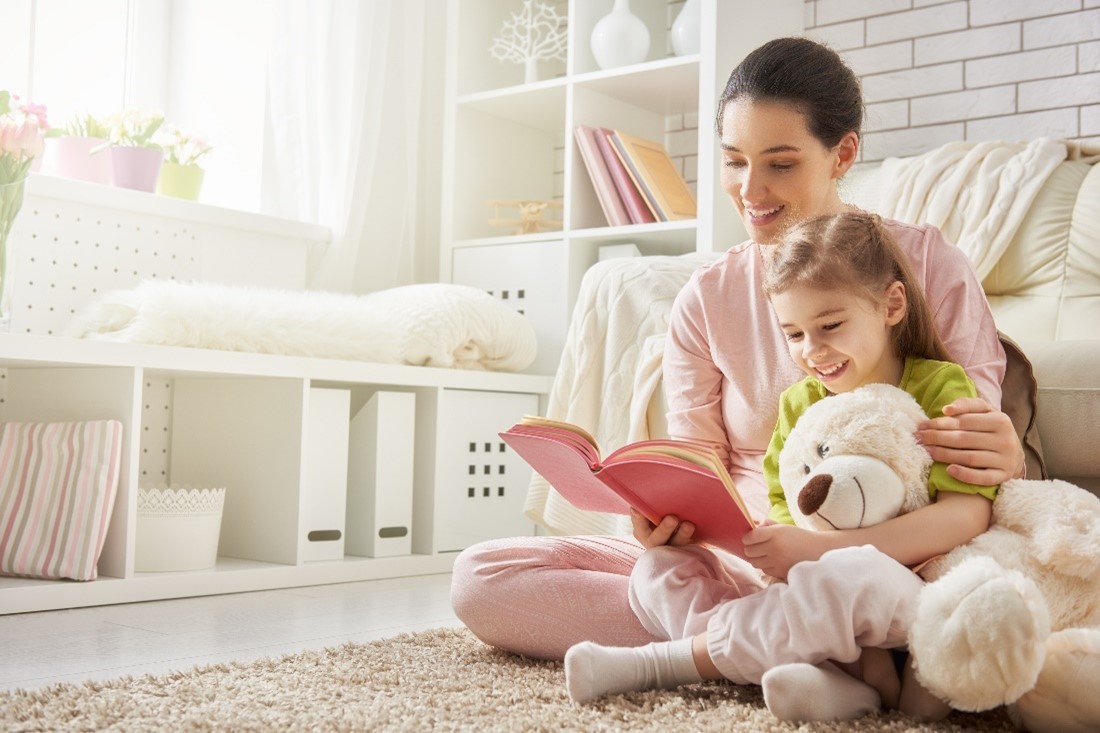 A Mother and Daughter Holding a Stuffed Bear Sit on a Fluffy Beige Rug Smiling and Reading a Book