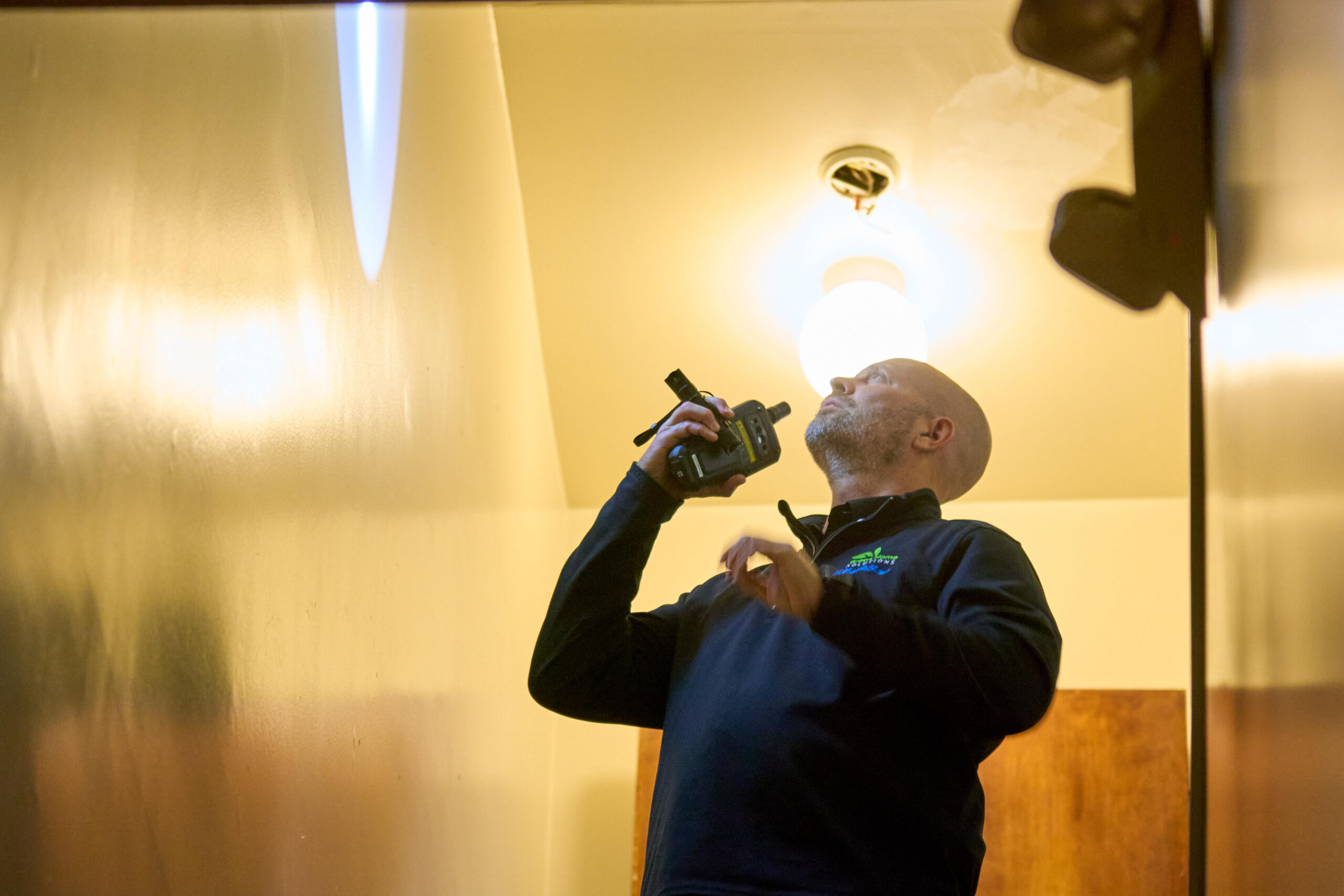 A Female Inspector in a Red Hard Hat Stands in a Stairway Hallway Holding a Pen and Clipboard and Looking Up at the Wall