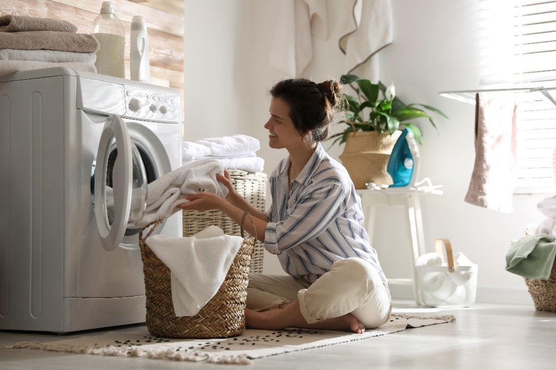 services5 A Smiling Woman in a Striped Shirt Sits on the Floor in Front of a Wicker Laundry Basket and Pulls Towels Out of a Dryer