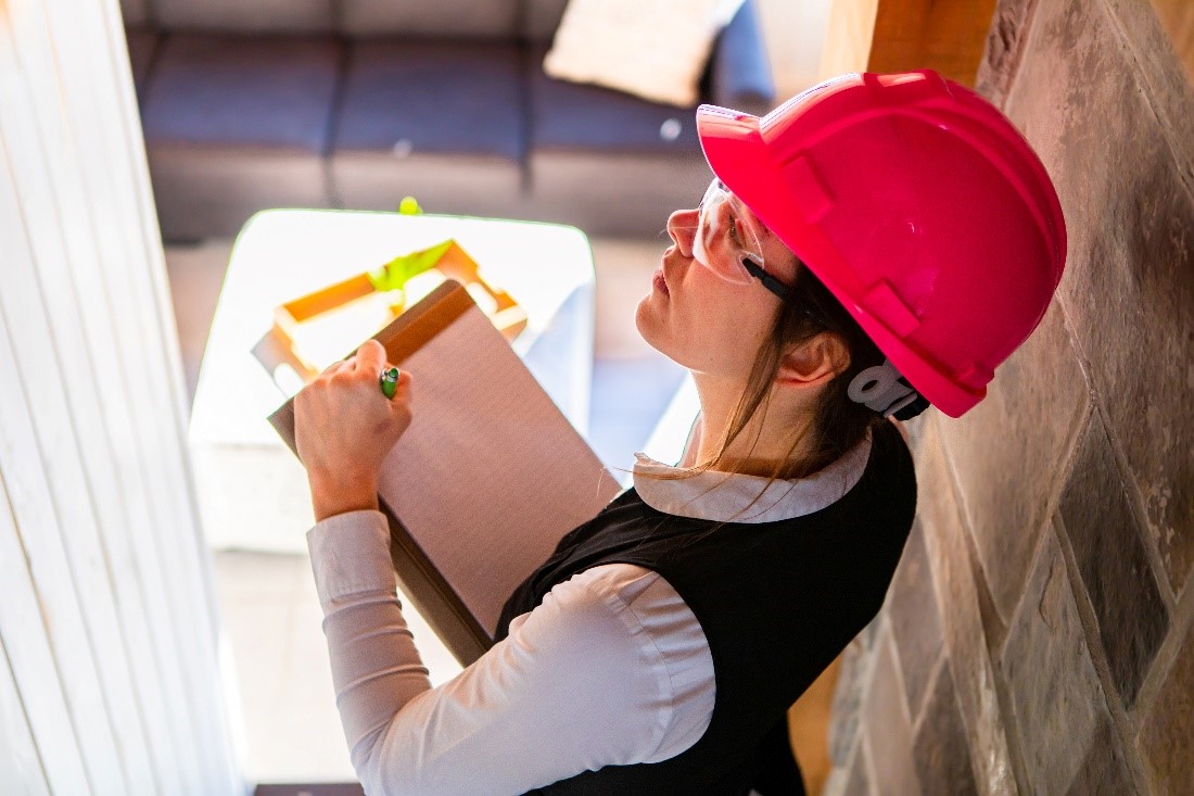 A Female Inspector in a Red Hard Hat Stands in a Stairway Hallway Holding a Pen and Clipboard and Looking Up at the Wall