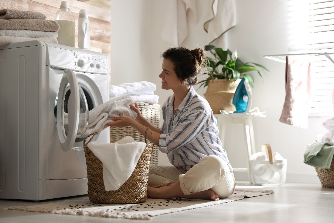A Smiling Woman in a Striped Shirt Sits on the Floor in Front of a Wicker Laundry Basket and Pulls Towels Out of a Dryer  