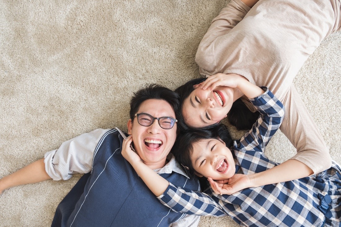 A Dad, Daughter, and Mom Lay on a Beige Carpet Smiling and Embracing Each Other‘s Faces