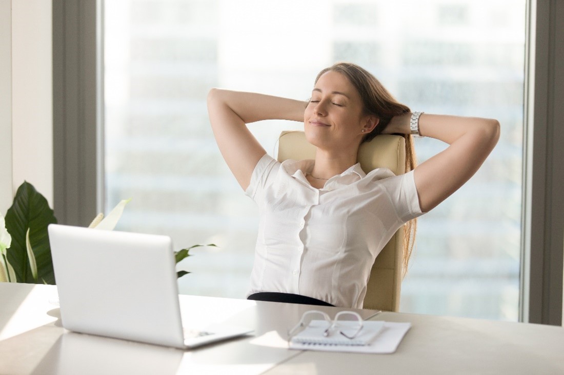 A Woman Sits at a Desk with an Open Laptop and Leans Back with Her Eyes Closed and Arms Behind Her Head Taking a Deep Breath     