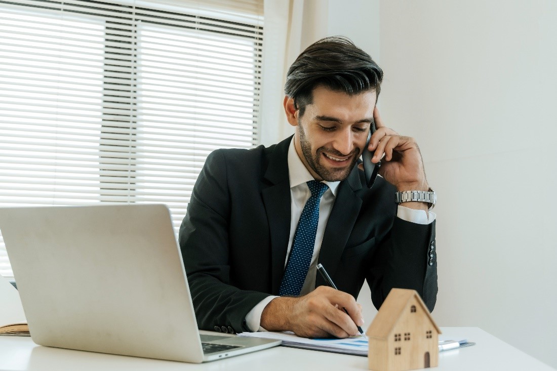 property-manager-inner A Man in a Business Suit at a Table with an Open Laptop and a House Figurine on it While on the Phone and Taking Notes