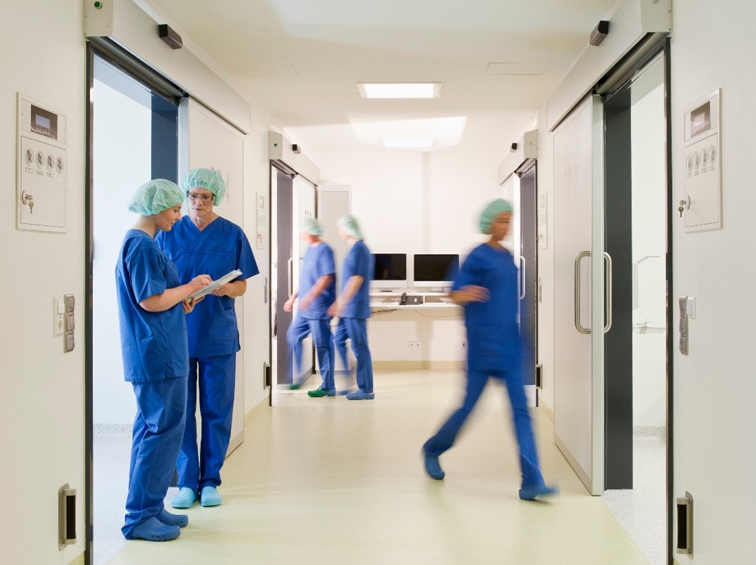 5 Healthcare Workers in Blue Scrubs and Hair Nets in Various Stages of Movement Through a Hospital Hallway 