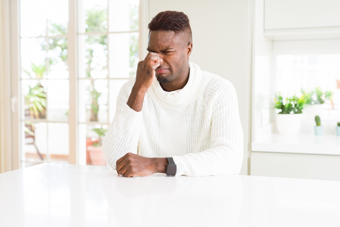 A Man in a White Turtleneck Sits at a Kitchen Counter and Plugs His Nose with a Facial Expression of Smelling Something Bad   