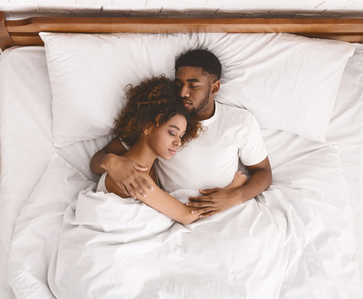 Picture1 A Young Adult Couple Peacefully Sleeping in an Embrace in White Bedding with a Wooden Headboard Behind Them