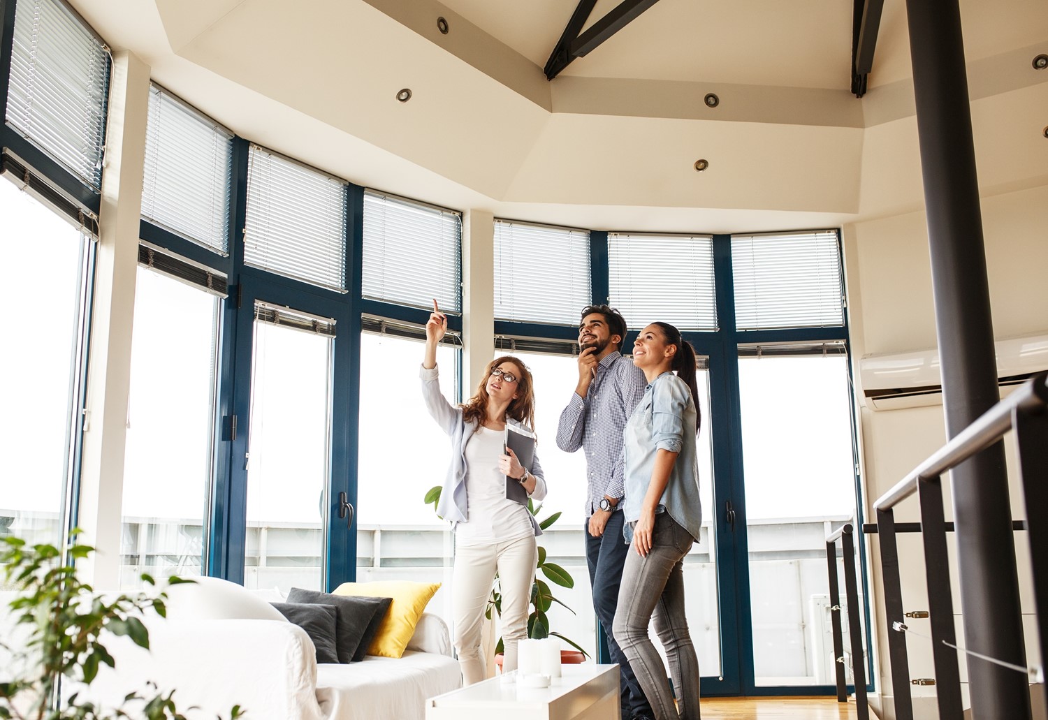 A Female Real Estate Agent Shows a Man and Woman the Vaulted Ceilings in a Room with Tall Windows and Recessed Lighting