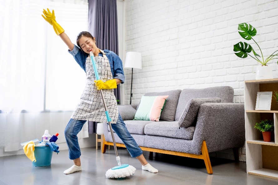 A woman spring cleaning her living room and using her mop as a microphone. 