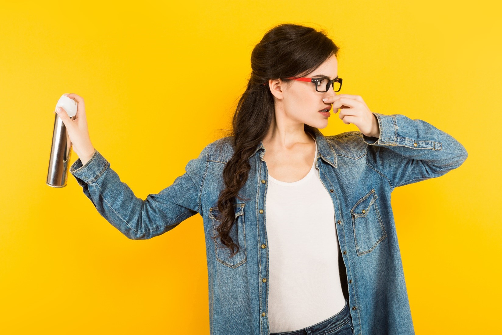 A woman wearing a denim jacket, holding her nose while spraying an air freshener in front of a yellow background.