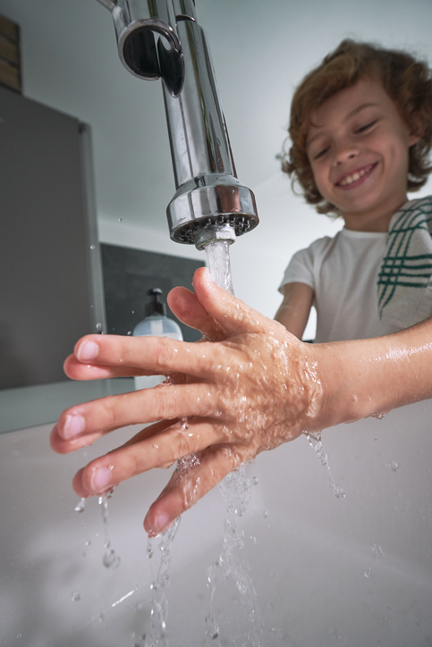 Content boy washing hands in bathroom Happy boy with towel washing hands under water in ceramic sink during daily hygienic procedure in light bathroom at home