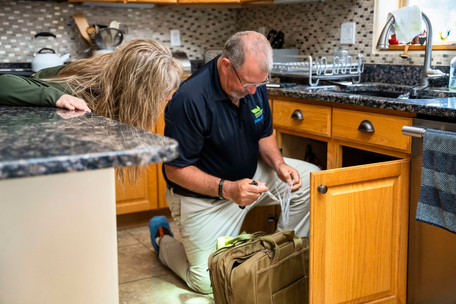 A man in a Green Home Solutions polo kneels on the floor and takes a sample of something from under a kitchen sink while a woman observes.