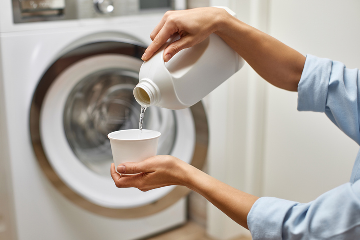 Woman pours liquid transparent laundry detergent or conditioner into plastic cap against washing machine.