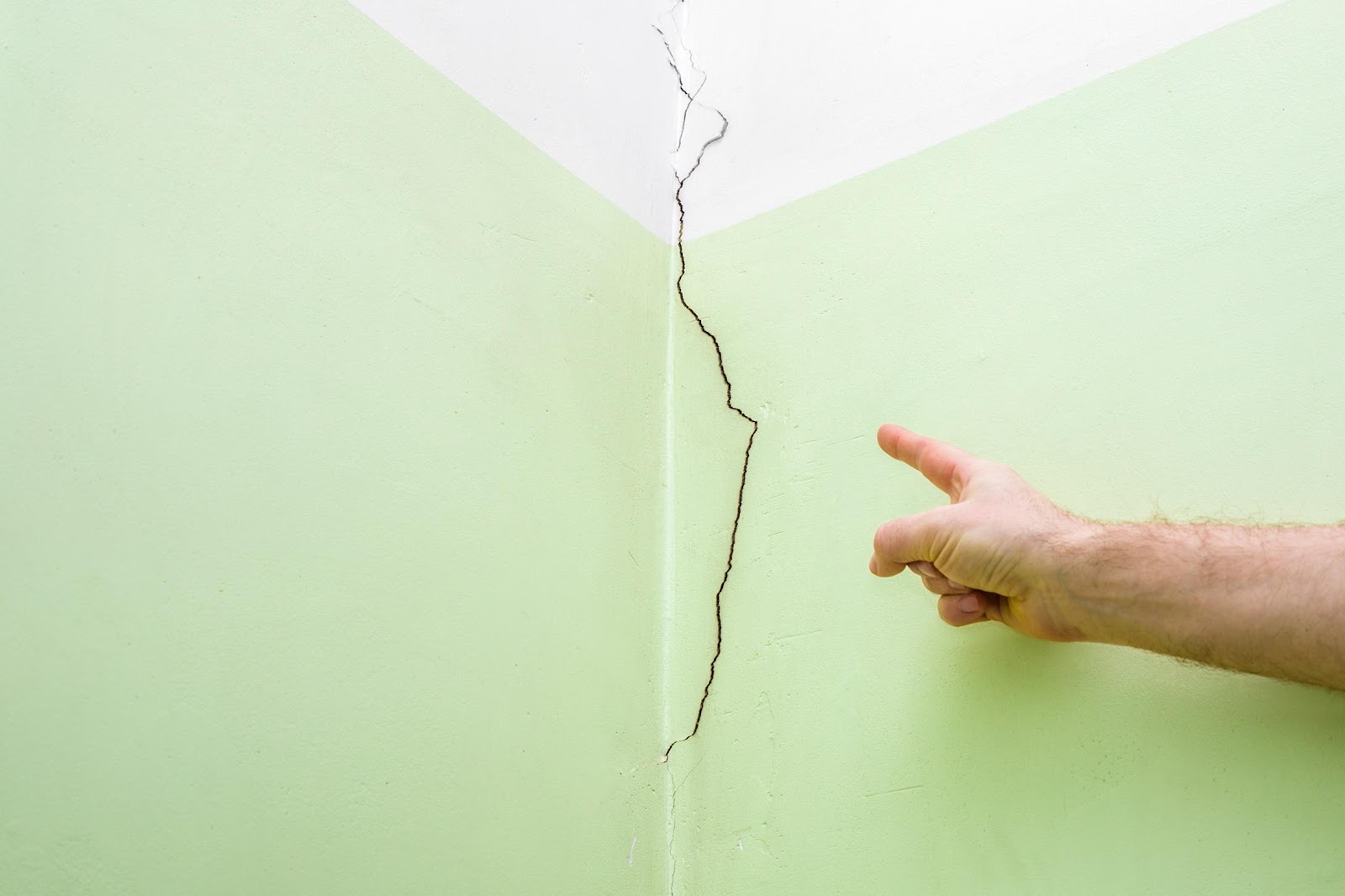 Foundation Crack Repair The corner of a room with a light green and white wall and a crack running down the wall near the corner with a man’s finger pointing at the crack.