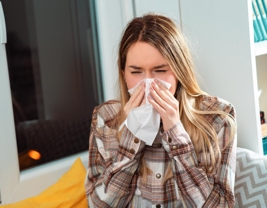 A woman wearing a plaid shirt and sitting in a chair next to a window and a bookshelf blows her nose into a tissue.