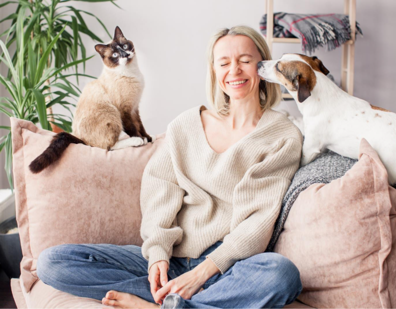A smiling woman sits on a couch with her eyes closed with a cat sitting on top a cushion on one side of her, and a dog on top of a cushion on the other side licking her face.